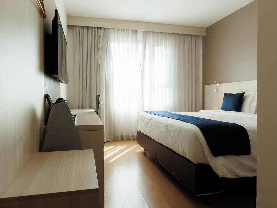 Habitación de hotel con cama, escritorio, silla, televisión y ventana con cortinas.
