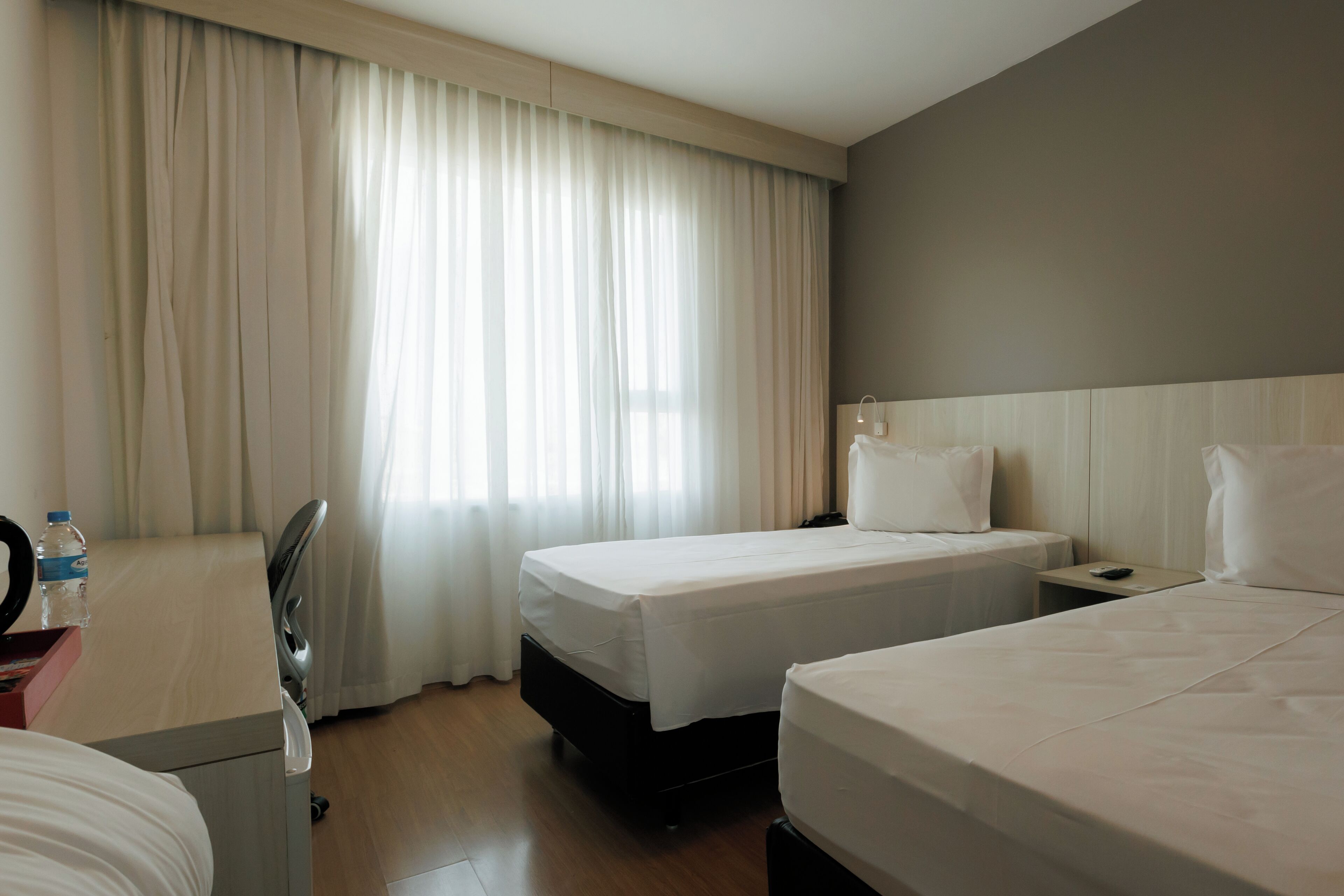 A hotel room with two beds, a desk with a chair, a mirror, and a window with curtains.