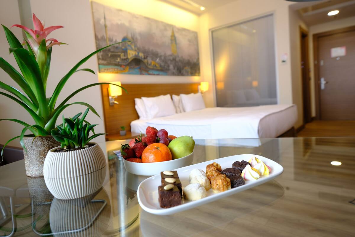 A hotel room with a bed, a painting on the wall, a fruit bowl, and a dessert tray on the table.