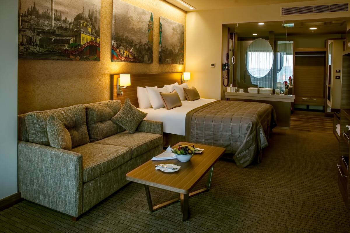 A hotel room with a bed, a sofa, a table with a bowl of fruit, and two wall-mounted lamps.