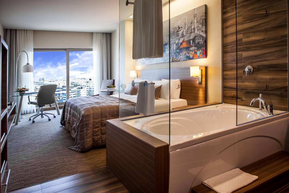 A modern hotel room with a large window, a private bathroom with a bathtub, a desk with a chair, and a bed with a view of the city.