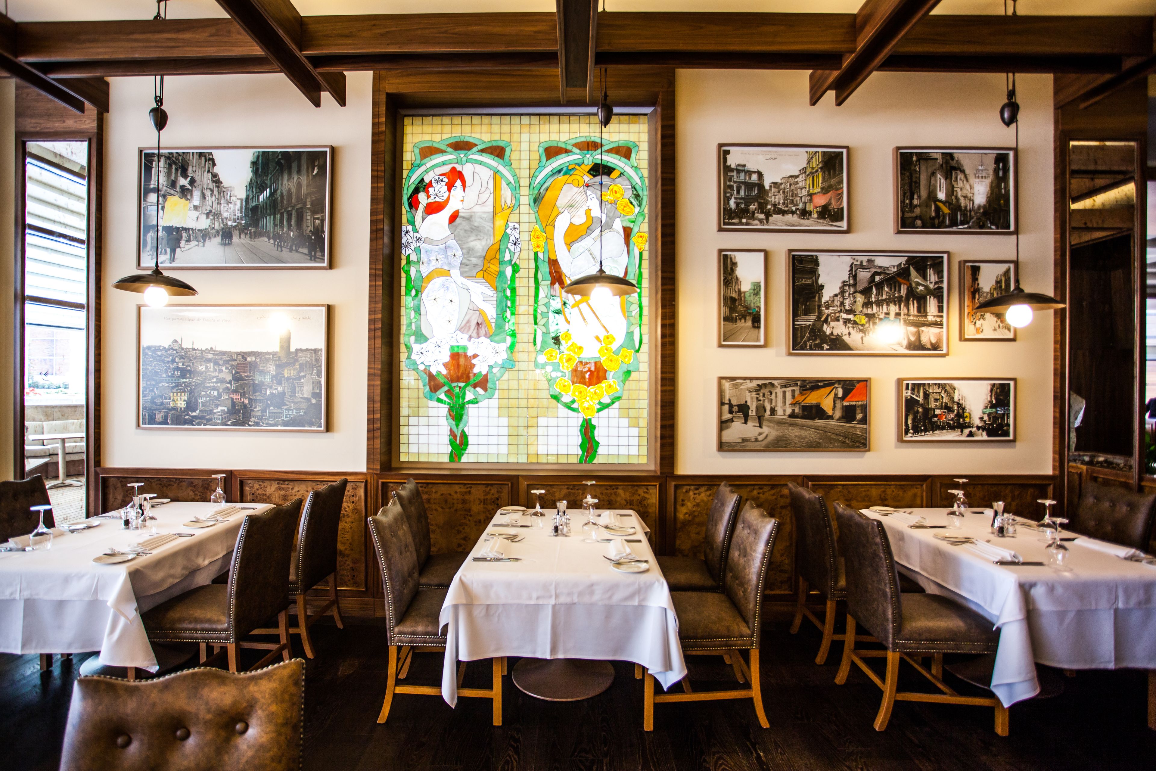 Restaurante com várias mesas preparadas para refeições, apresentando uma janela de vitrais e fotografias emolduradas nas paredes.
