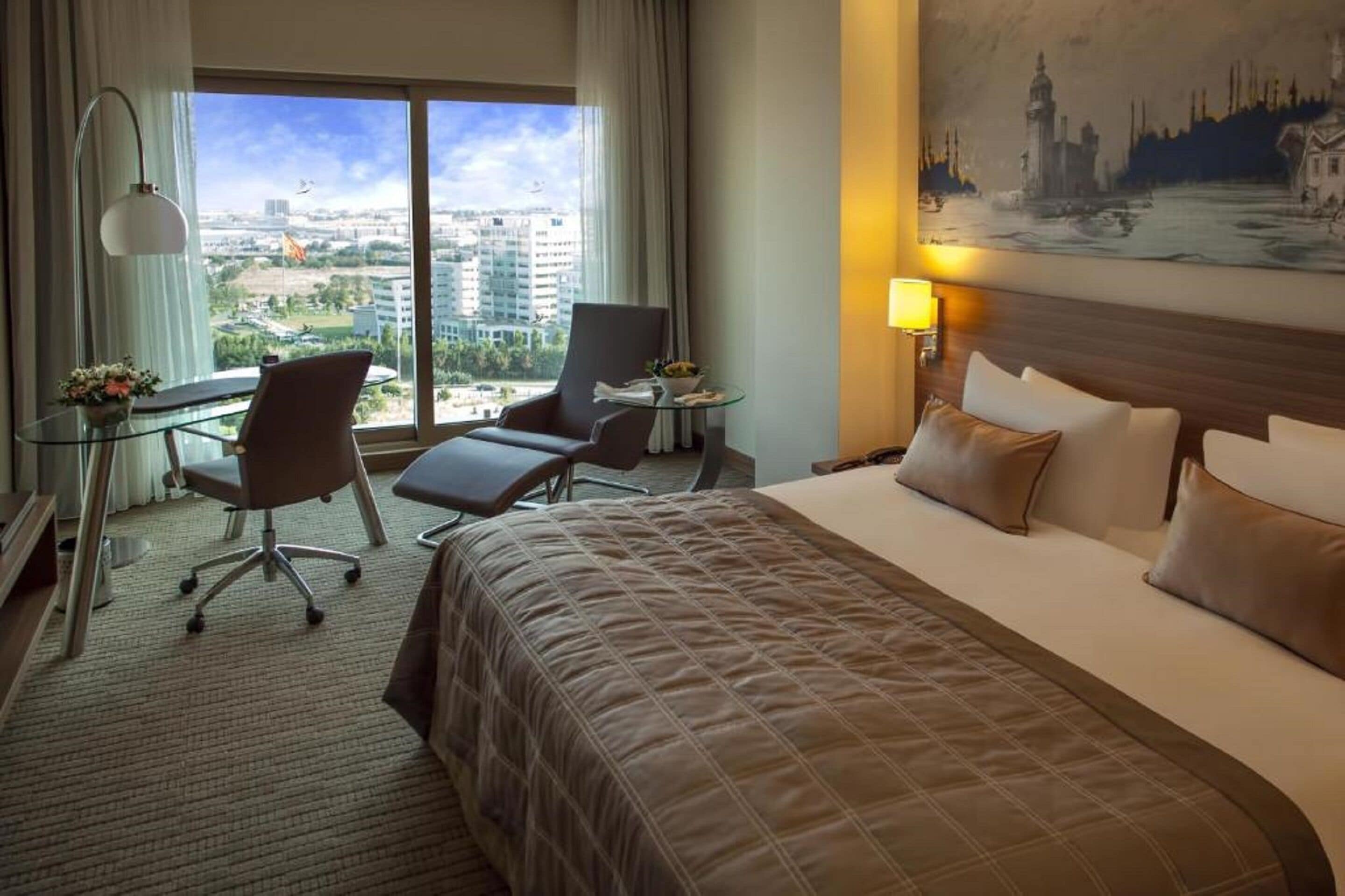 Une chambre d’hôtel avec un grand lit, deux fauteuils, un bureau avec une lampe et une vue sur le paysage urbain grâce à de grandes fenêtres.