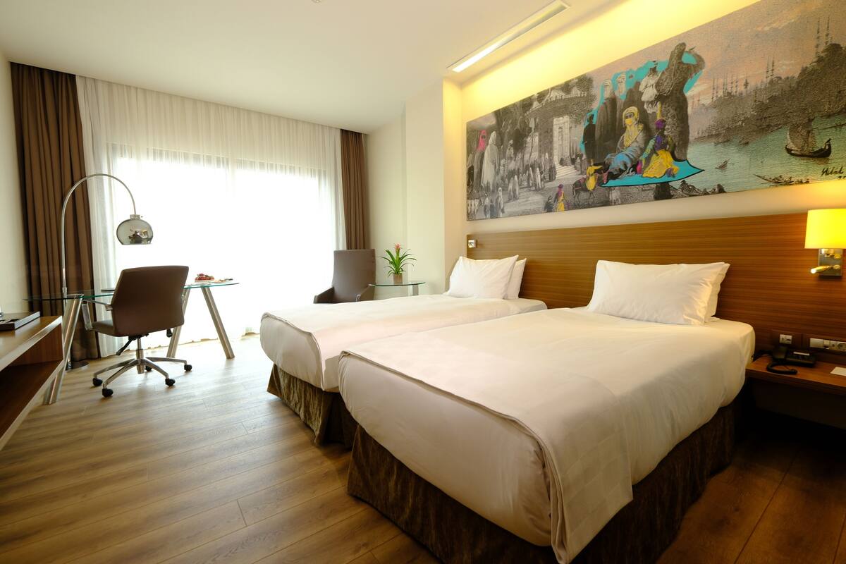 A hotel room with two beds, a desk with a chair, a lamp, and a painting on the wall.