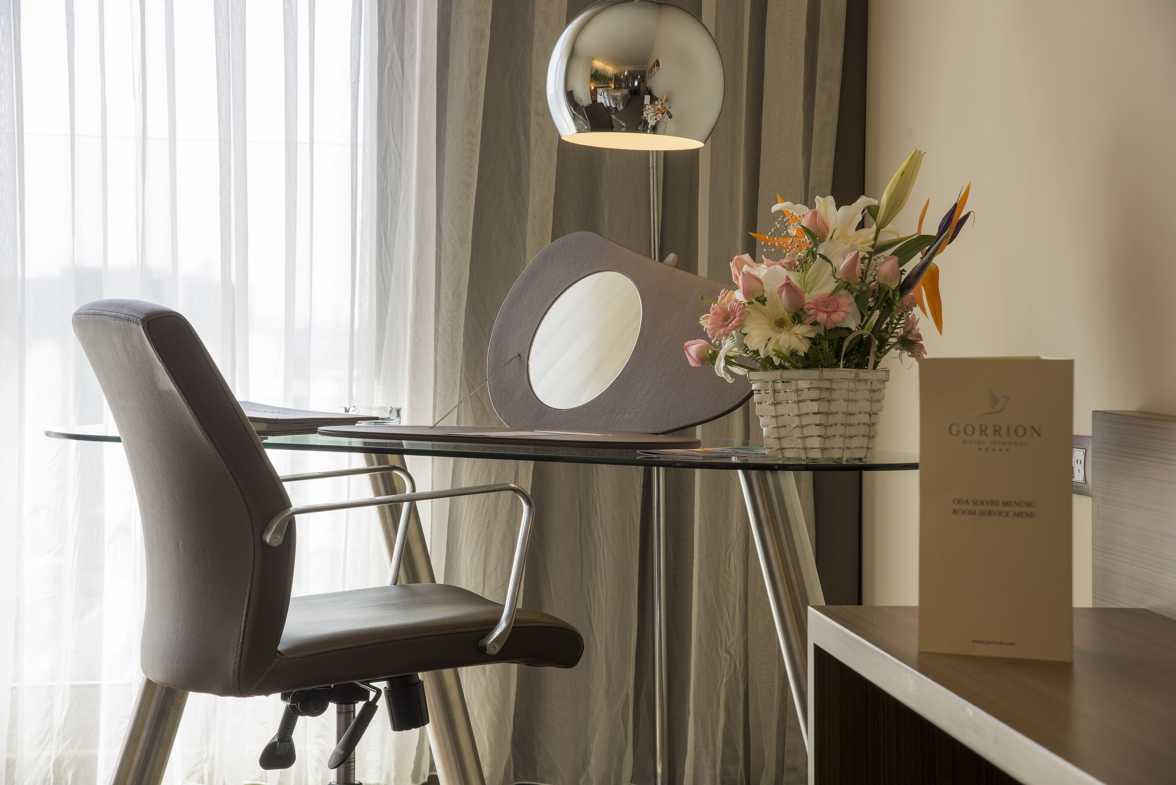 Une chambre d’hôtel moderne avec un bureau en verre, une chaise, un miroir décoratif, un vase avec des fleurs et le menu du service d’étage Gorrion.
