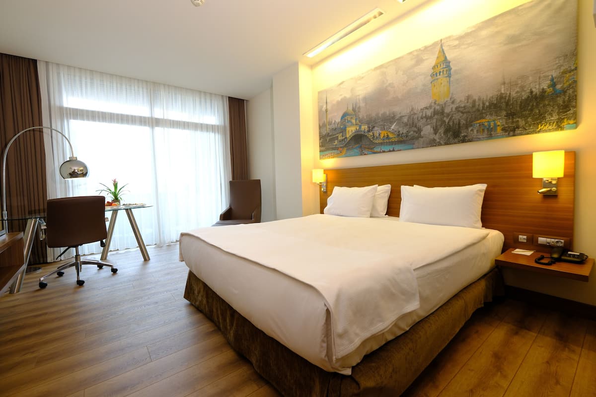 A hotel room with a large bed, a desk, a chair, and a painting of a tower on the wall.