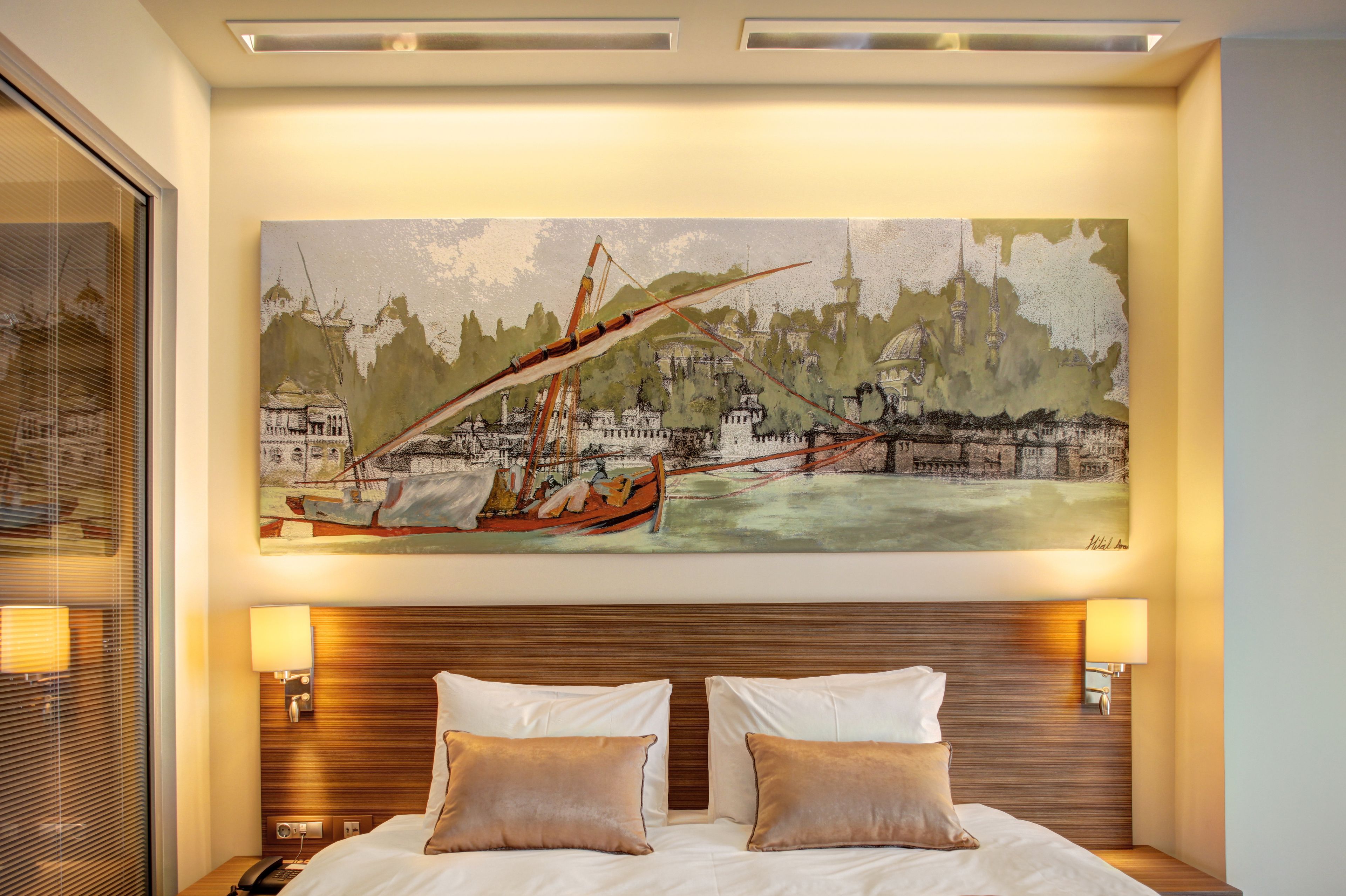 A hotel room with a large painting of a boat on a river, a wooden headboard, two bedside lamps, and a nightstand with a phone.