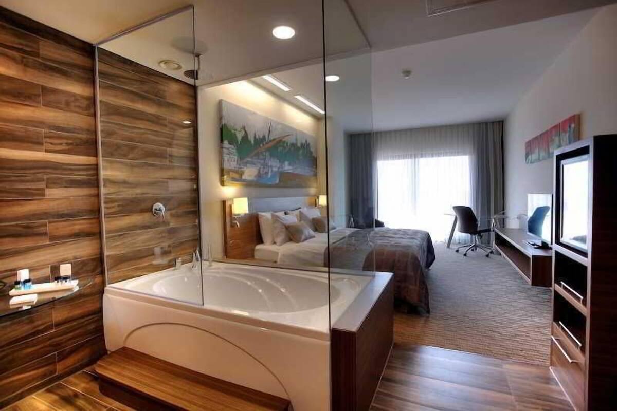 A modern hotel room with a large bathtub, a bed, a desk, and a television.