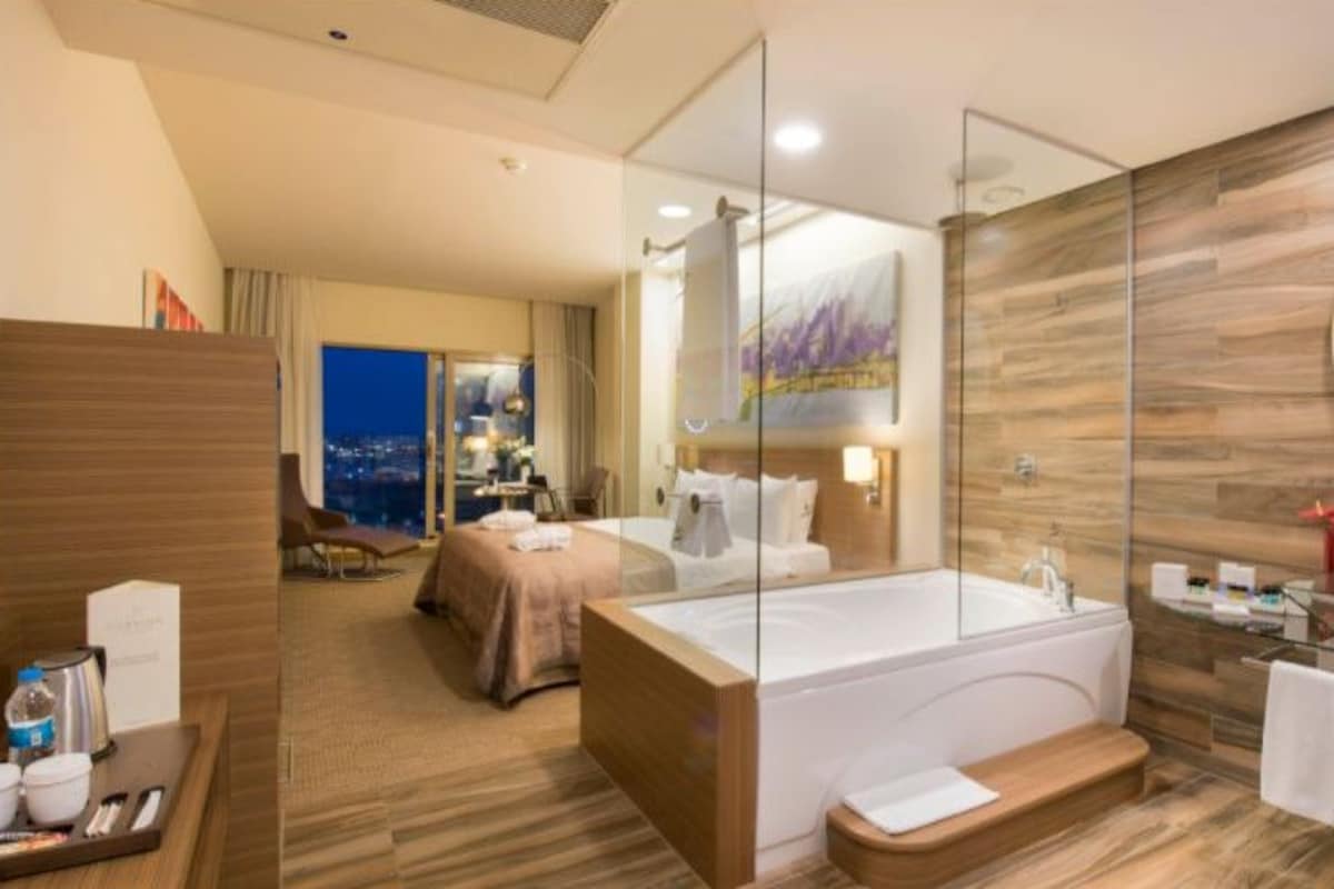 A modern hotel room with a large bathtub, a separate bedroom with a view, and a glass shower enclosure.