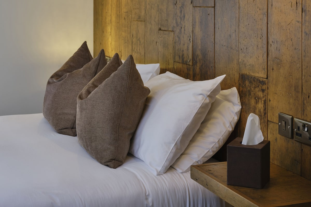Een net opgemaakt bed met witte lakens en bruine kussens, een nachtkastje met een zakdoosje en twee wandlampen.