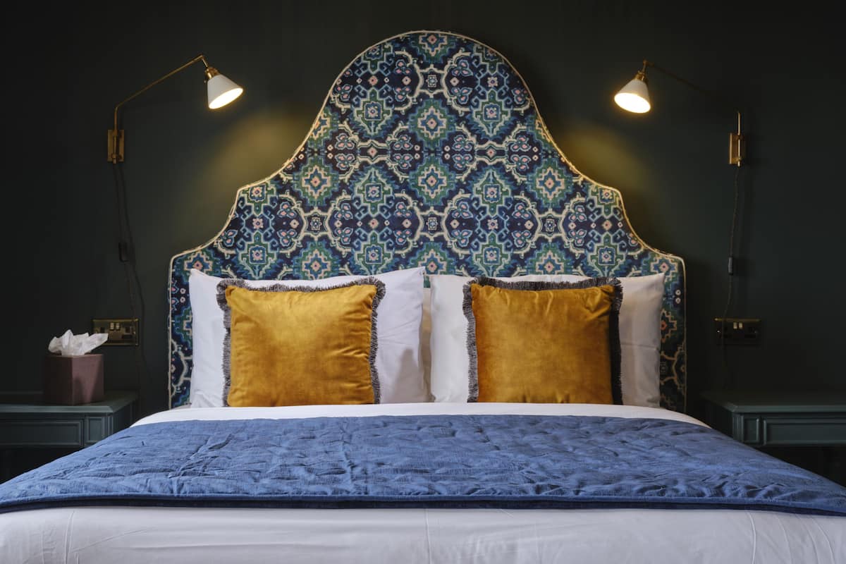Een bed met een hoofdbord met een patroon, twee bedlampen en een blauwe deken.
