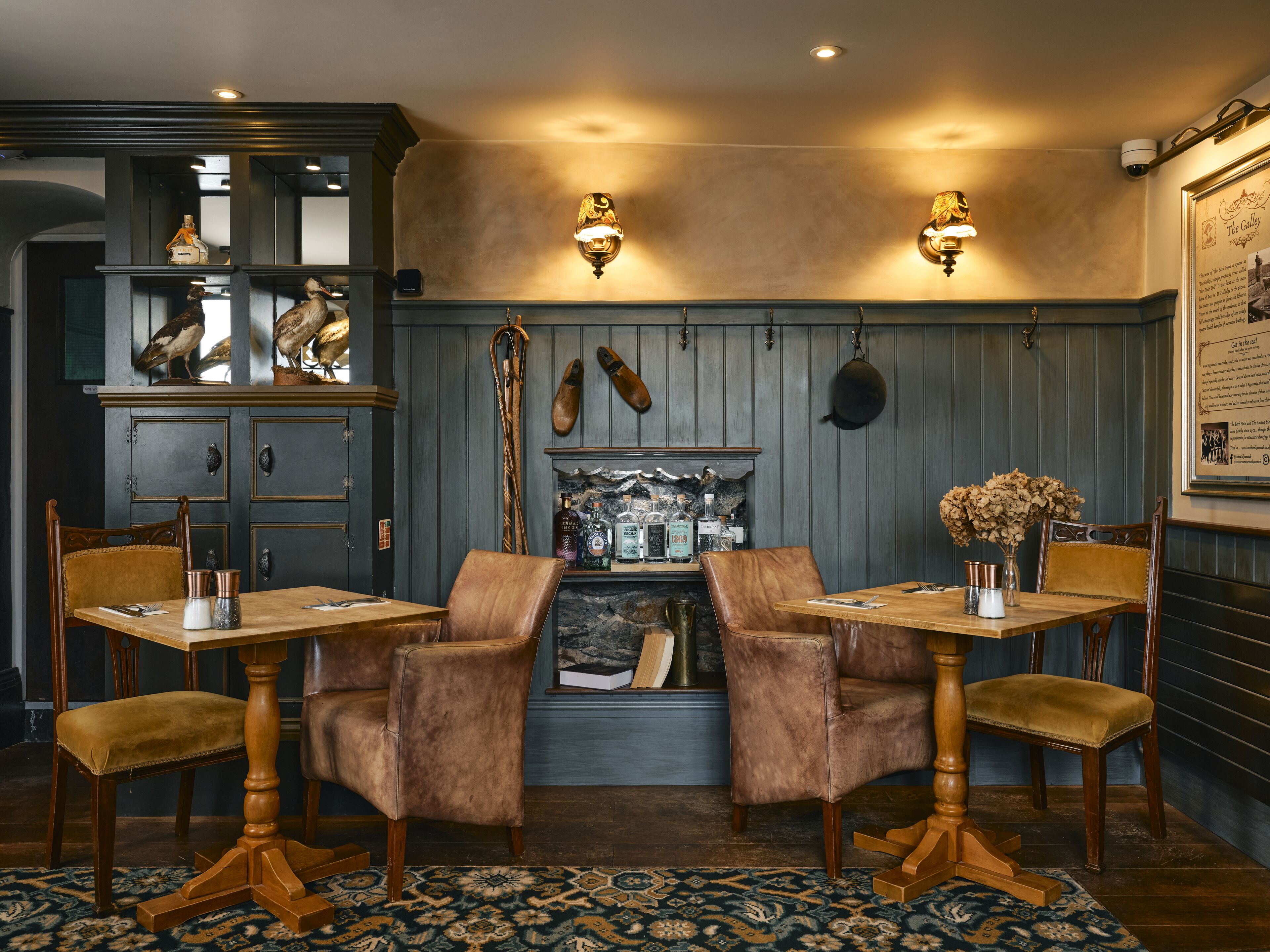 Un espace repas avec des tables et des chaises en bois, une cheminée avec un manteau en pierre et des luminaires fixés au mur.