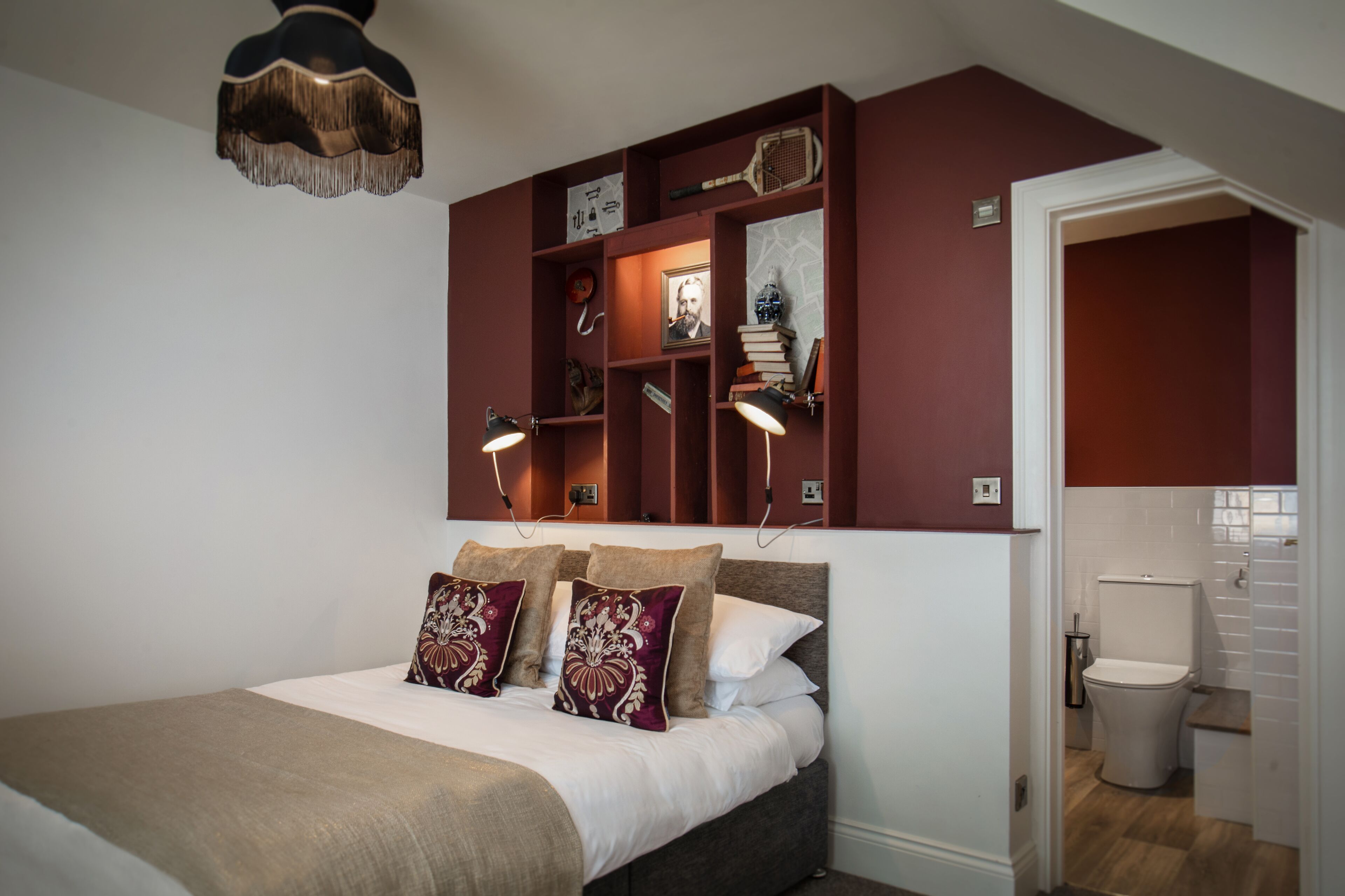 A bedroom with a bed, bedside lamps, a shelf with books and decorative items, and a view of the bathroom.