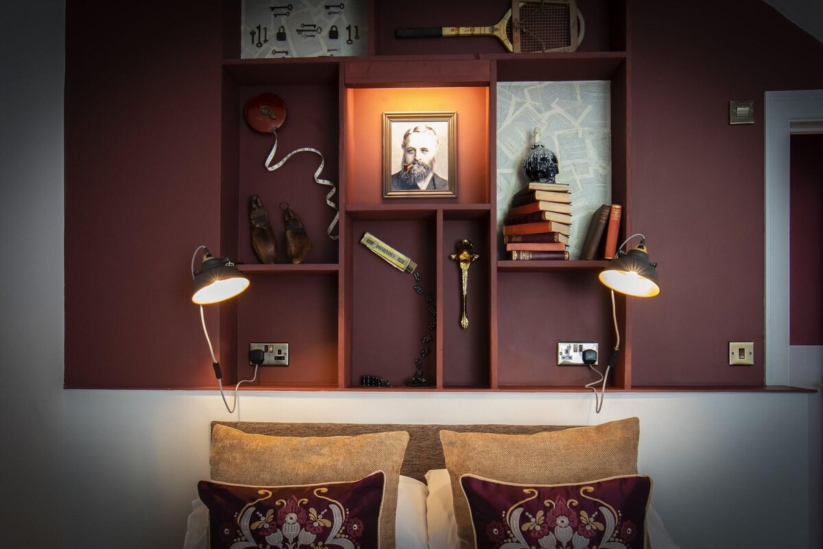 Een kamer met een donkerrode wand, een houten plank met diverse spullen, twee lampen, en een bed met gekleurde kussens.