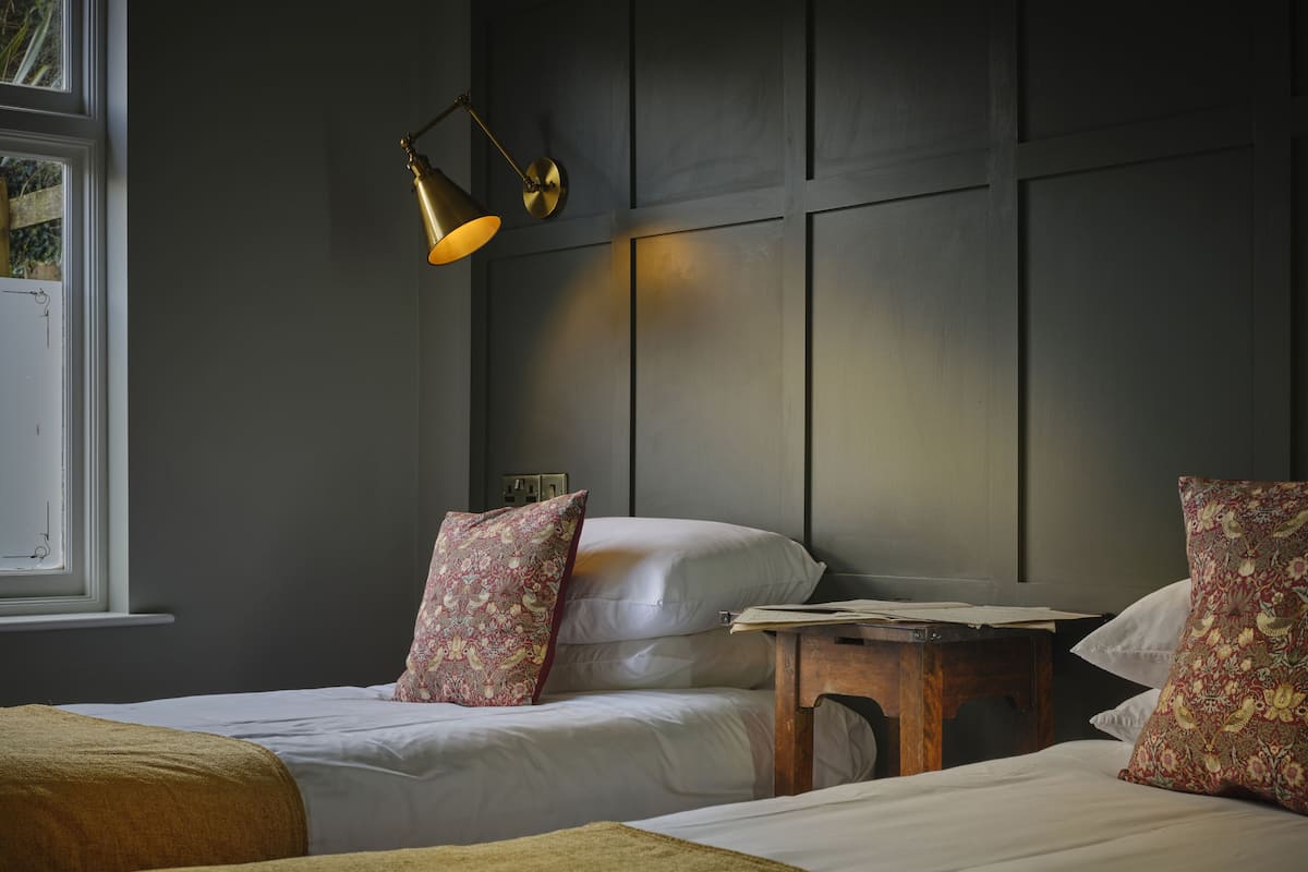 Een hotelkamer met twee bedden, een houten nachtkastje, een wandlamp en een raam met uitzicht op groen.
