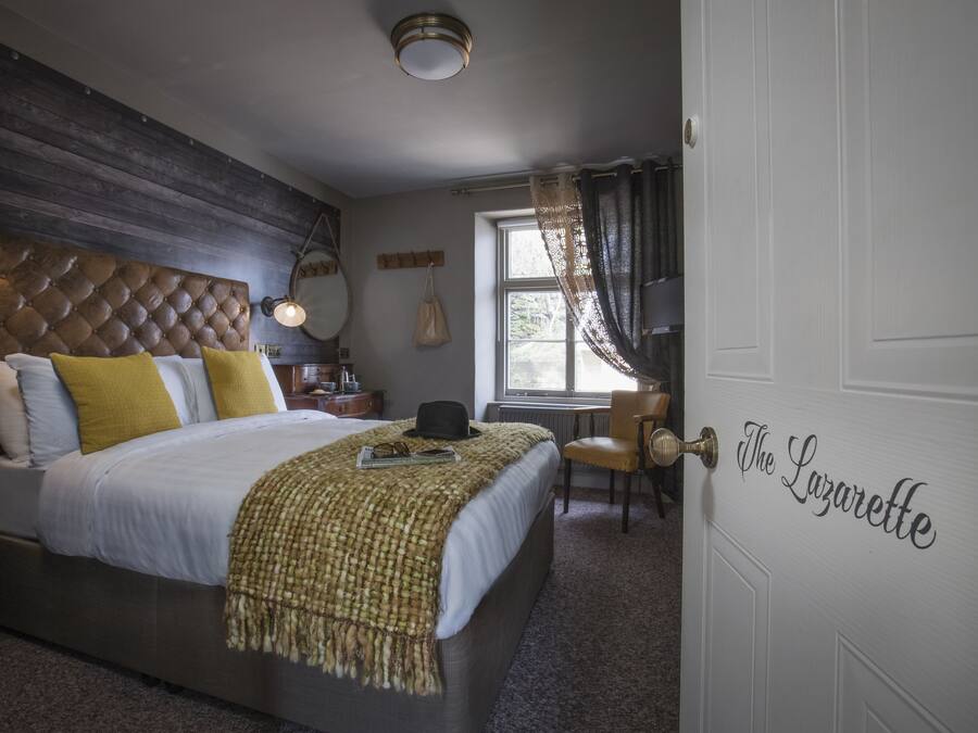 A bedroom with a tufted headboard, a bed with white and yellow bedding, a bedside table with a lamp, and a window with curtains.