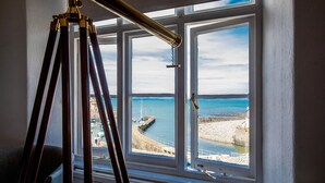 Une pièce dotée d’une grande fenêtre donnant sur une zone côtière, un trépied, et une vue sur la mer et la plage.