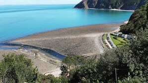Un paysage côtier avec une plage de galets, une mer bleue et limpide, et un petit village composé de maisons et d’une route.