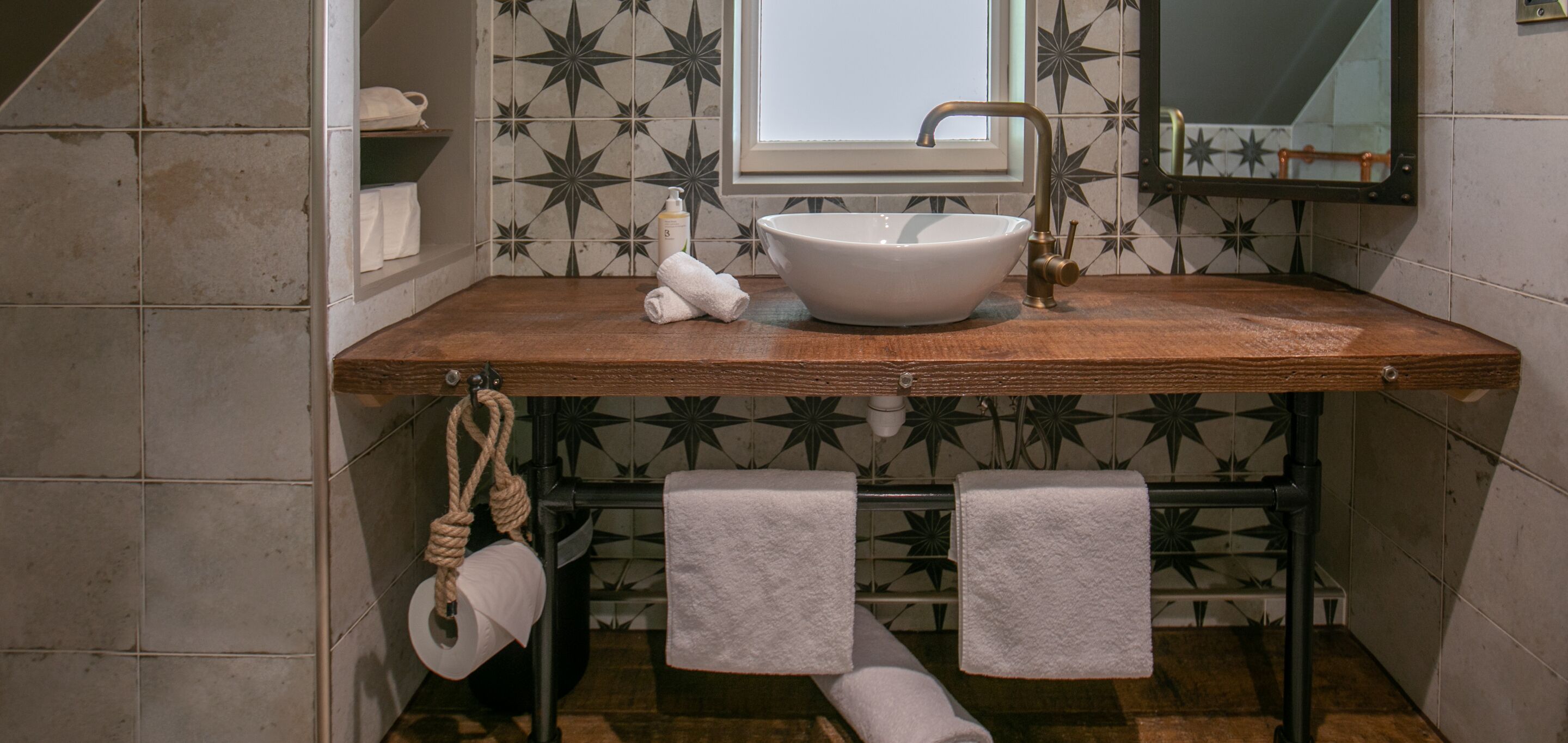 A bathroom with a wooden vanity, a white sink, and a mirror. There are two towels hanging on hooks and a toilet paper roll on a rope.
