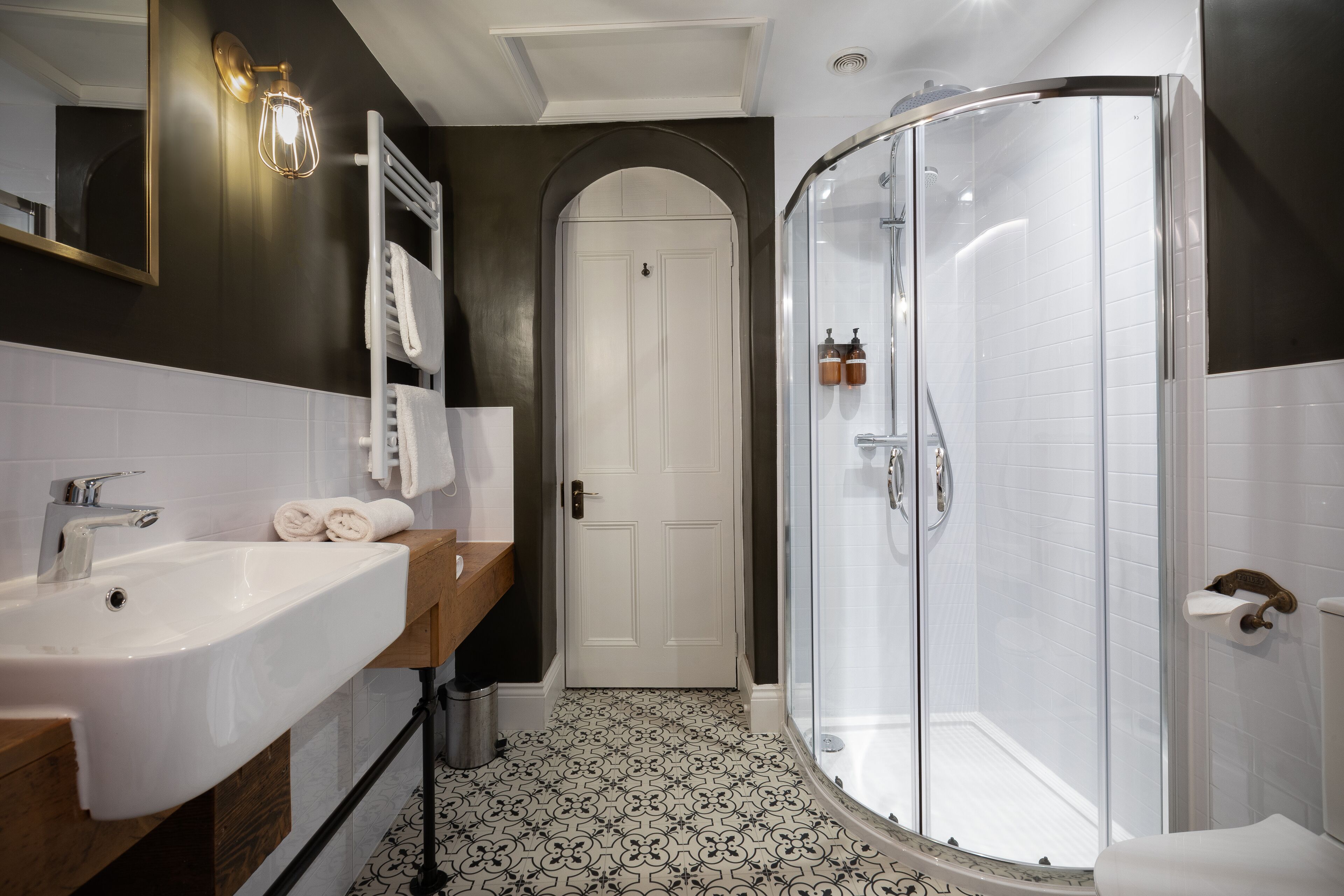 Ein Badezimmer mit einem weißen Waschbecken, einem Spiegel, einem Handtuchhalter, einer Dusche mit Glastür und einem gemusterten Boden.