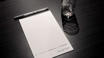 Una libreta rayada con la etiqueta "thought pad", un bolígrafo y un vaso de agua sobre una mesa de madera oscura.