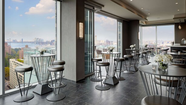 A modern bar area with high tables and stools, large windows offering a city view, and a minimalist interior design. - Hampton Inn Brooklyn/Downtown (Brooklyn)