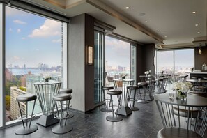 A modern bar area with high tables and stools, large windows offering a city view, and a minimalist interior design. - Hampton Inn Brooklyn/Downtown (Brooklyn)