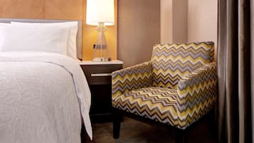 A hotel room with a bed, a nightstand with a lamp, and a chair with a zigzag pattern.
