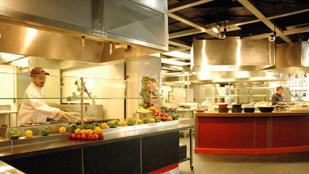 A commercial kitchen with stainless steel appliances, a central island with fruits and vegetables, and two chefs working. - University of Toronto - Wilson Hall Residence (Toronto)