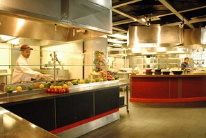 A commercial kitchen with stainless steel appliances, a central island with fruits and vegetables, and two chefs working. - University of Toronto - Wilson Hall Residence (Toronto)