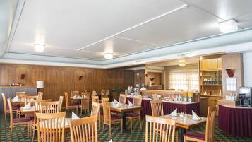 Comedor de hotel con varias mesas y sillas, una cocina al fondo y una alfombra estampada.
