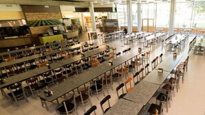 Eine Cafeteria mit mehreren langen Tischen und Stühlen, einem Küchenbereich und großen Fenstern, die viel Tageslicht hereinlassen.