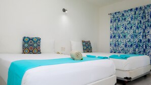 Une chambre d’hôtel avec deux lits, du linge de lit blanc, une couverture bleue, des coussins à motifs et un rideau à fleurs.