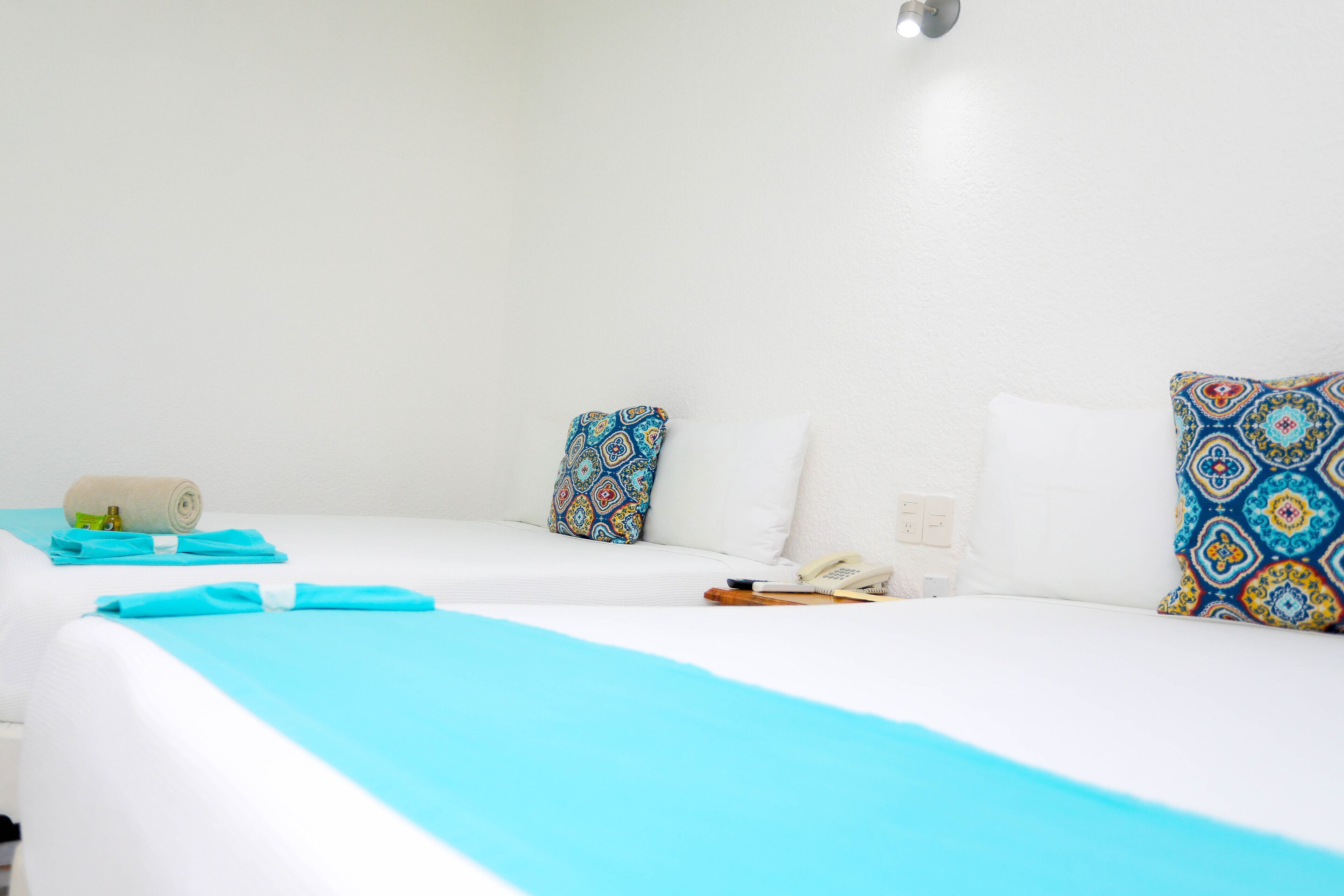 A hotel room with two beds, each with a white mattress and blue bedding. There are patterned pillows on the beds, a rolled towel on the left bed, and a telephone on the right bed.