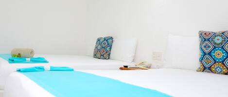 A hotel room with two beds, each with a white mattress and blue bedding. There are patterned pillows on the beds, a rolled towel on the left bed, and a telephone on the right bed.