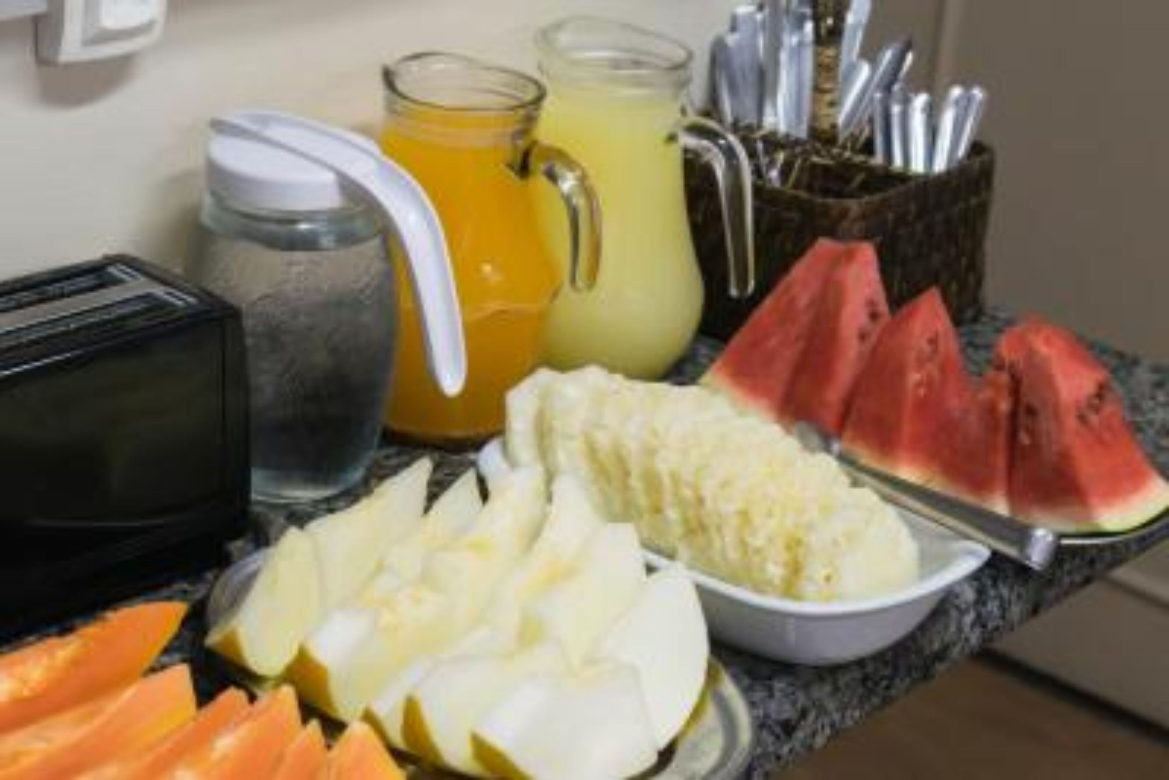 A breakfast spread with sliced fruits, a bowl of cereal, and two pitchers of juice on a kitchen counter.