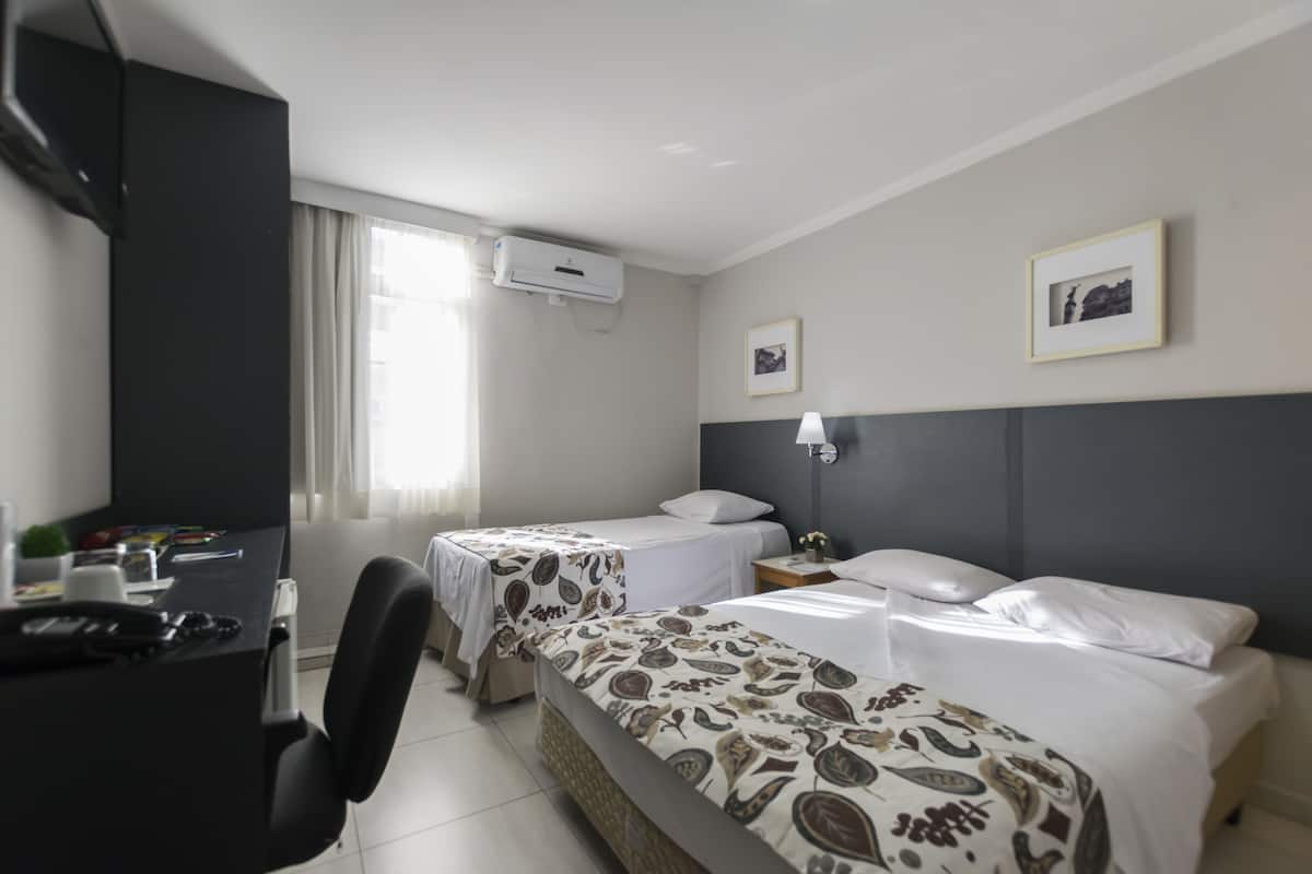 Une chambre d’hôtel avec deux lits, un bureau, une chaise, une table de chevet, une lampe et une fenêtre avec des rideaux.