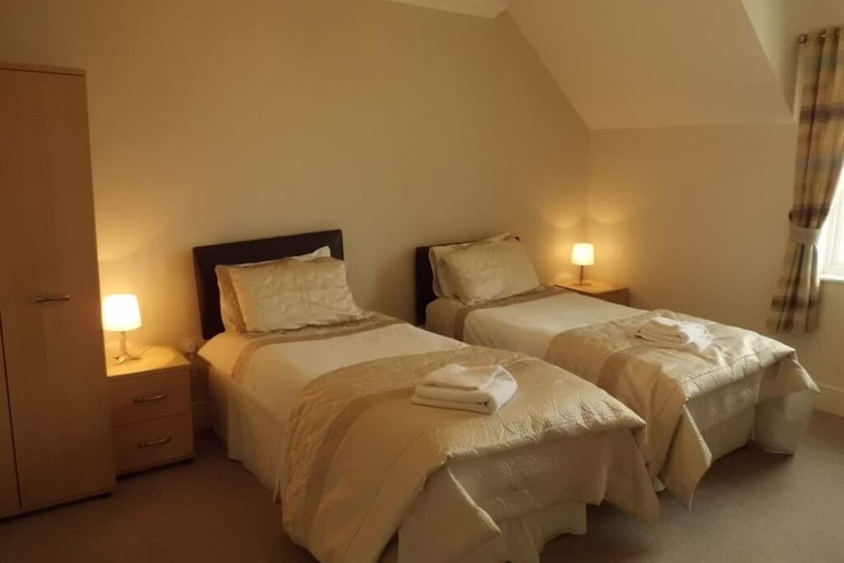 A room with two beds, each with a bedside table and lamp, a wardrobe, and a window with curtains.