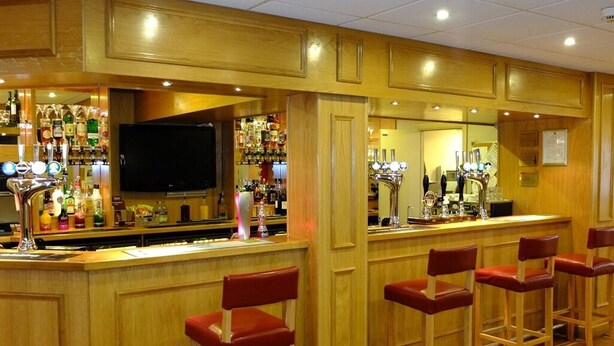 Zona de bar con paneles de madera, un televisor y una variedad de botellas y vasos en estanterías.