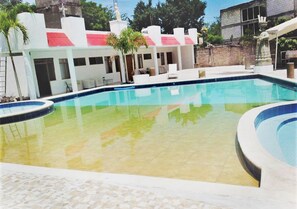 A swimming pool with a tiled deck, a covered seating area, and a building with a red roof in the background.