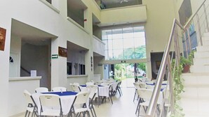 Un espacio interior amplio con varias mesas y sillas, una escalera y ventanas grandes que dejan entrar luz natural.