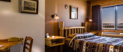A cozy room with a wooden bed, a desk, and a chair. There are framed pictures on the wall and a window with a view.