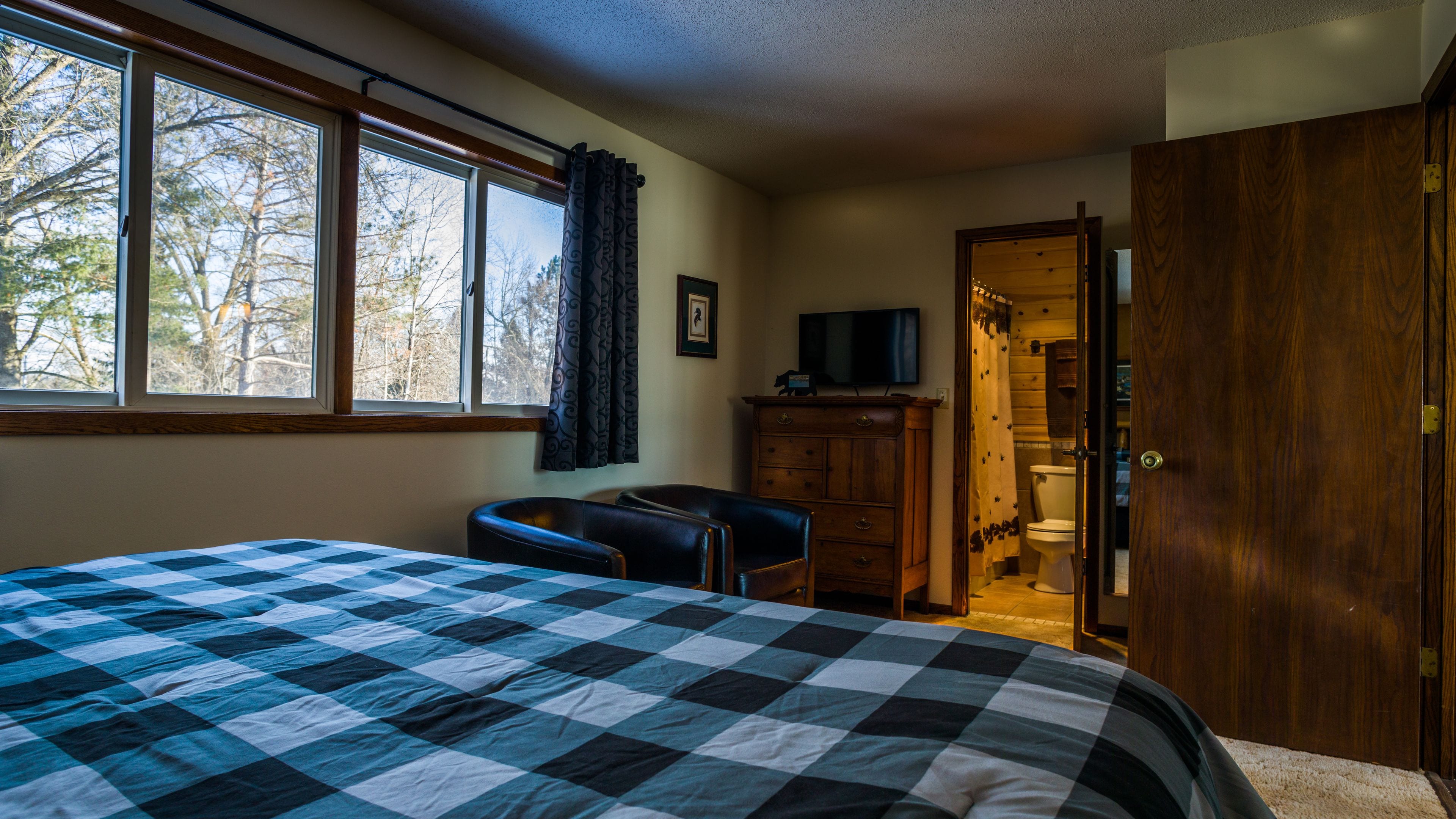 A bedroom with a large bed, a desk, a chair, a wooden dresser, and a bathroom visible through an open door.