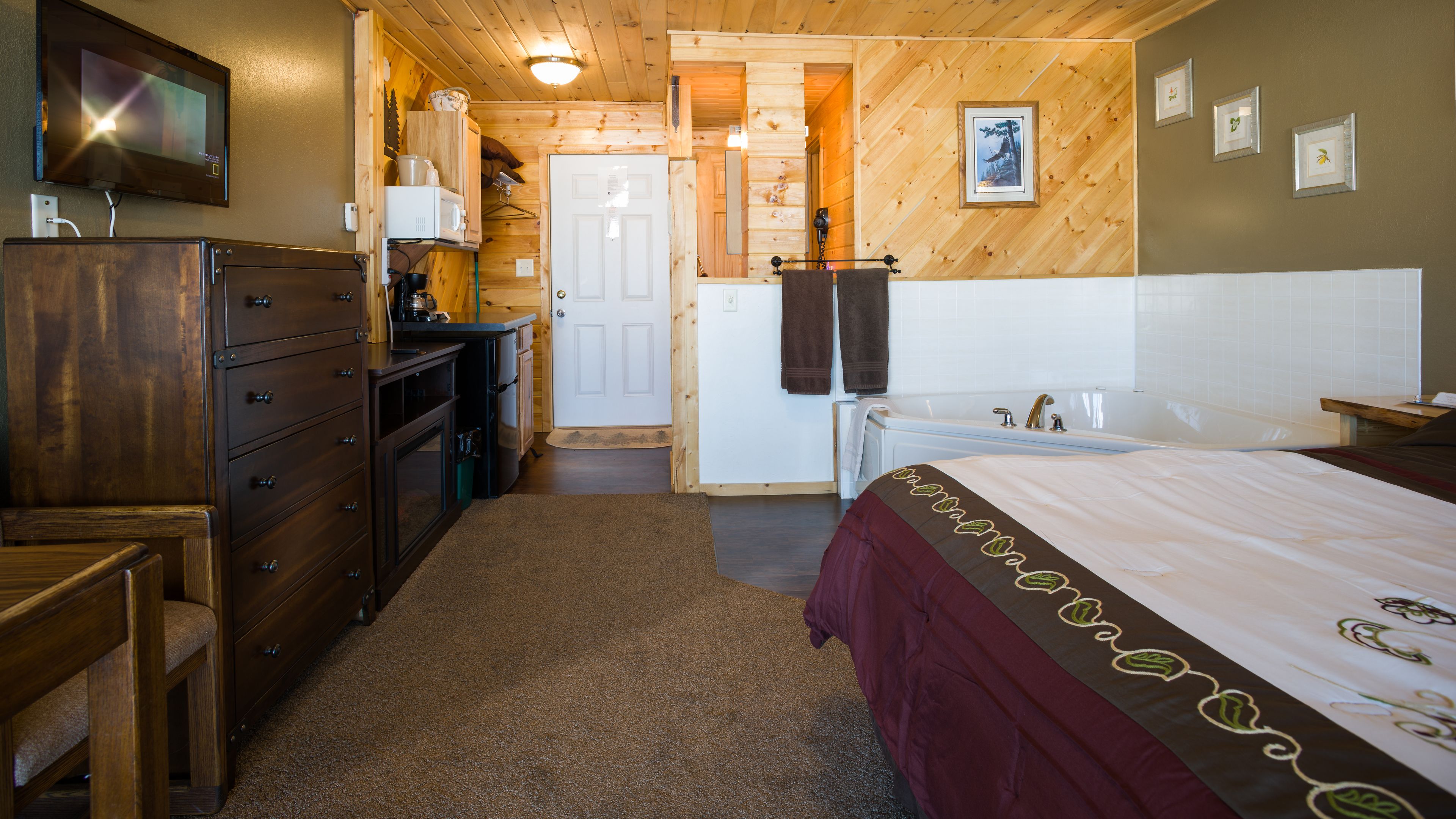 Una habitación acogedora con suelo de madera, una cama con colcha roja y blanca, un aparador, un televisor y una puerta que da a otra habitación.
