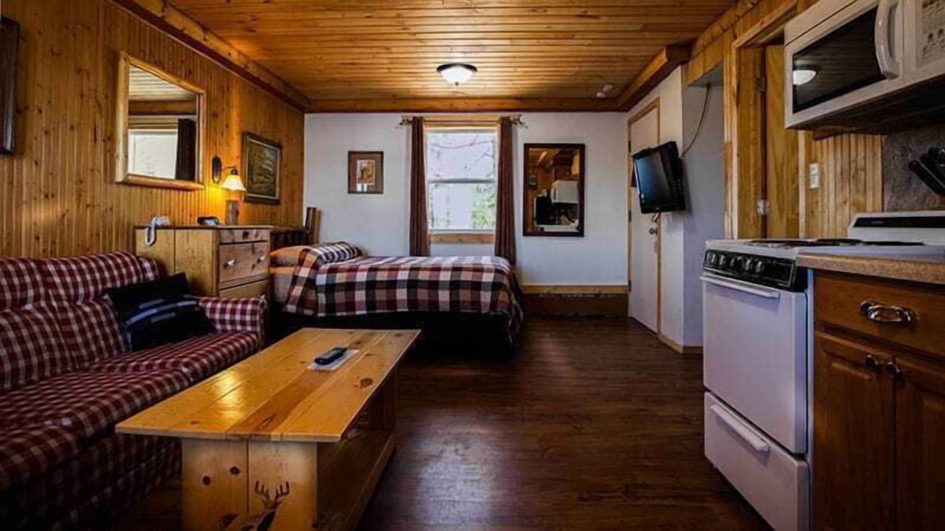 A cabin-style room with a wooden interior, a sofa, a bed, a dining table, a TV, and a kitchen area with a stove.