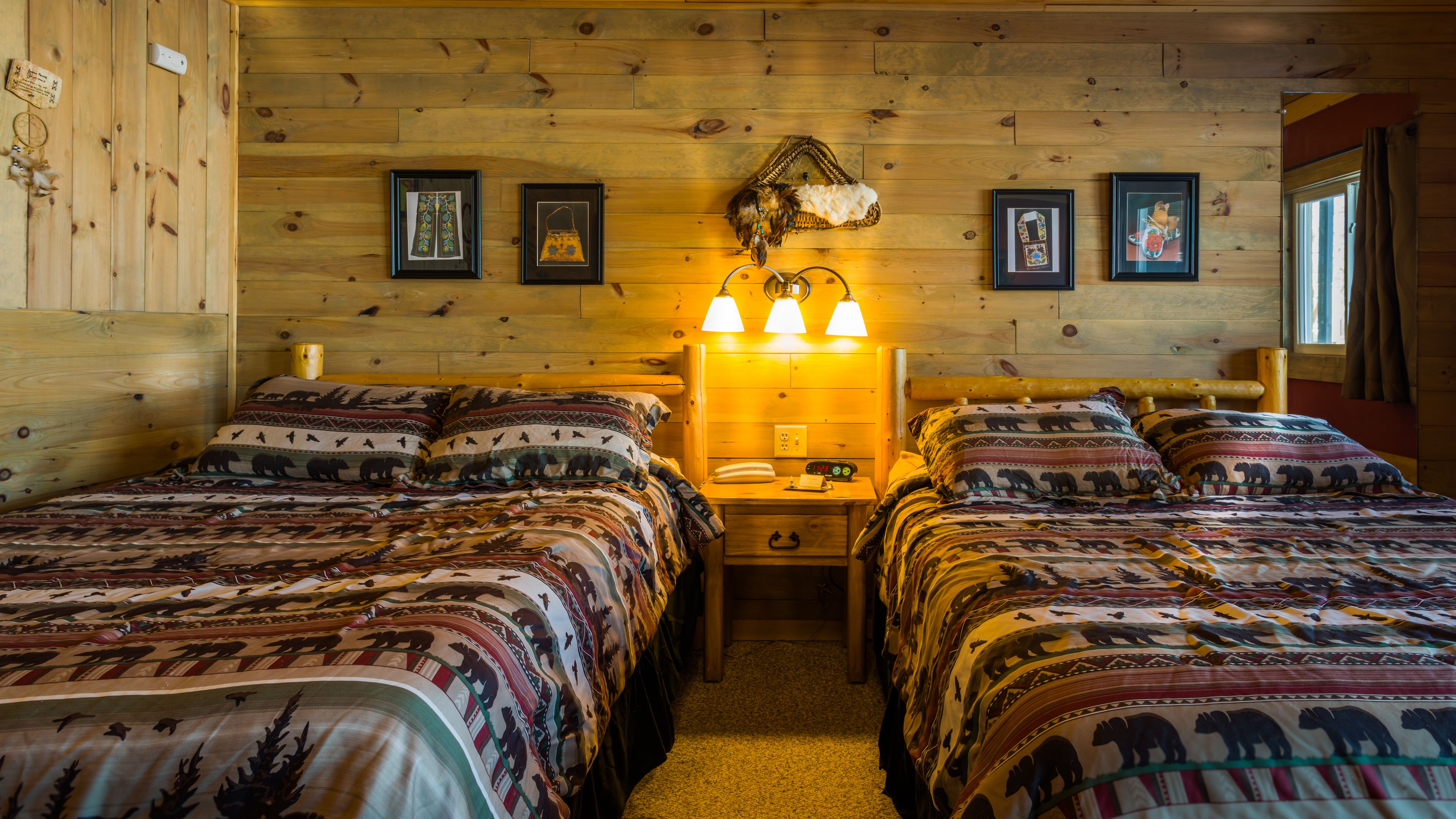 A wooden cabin room with two beds, each with patterned bedding, a nightstand with a lamp, and framed pictures on the walls.