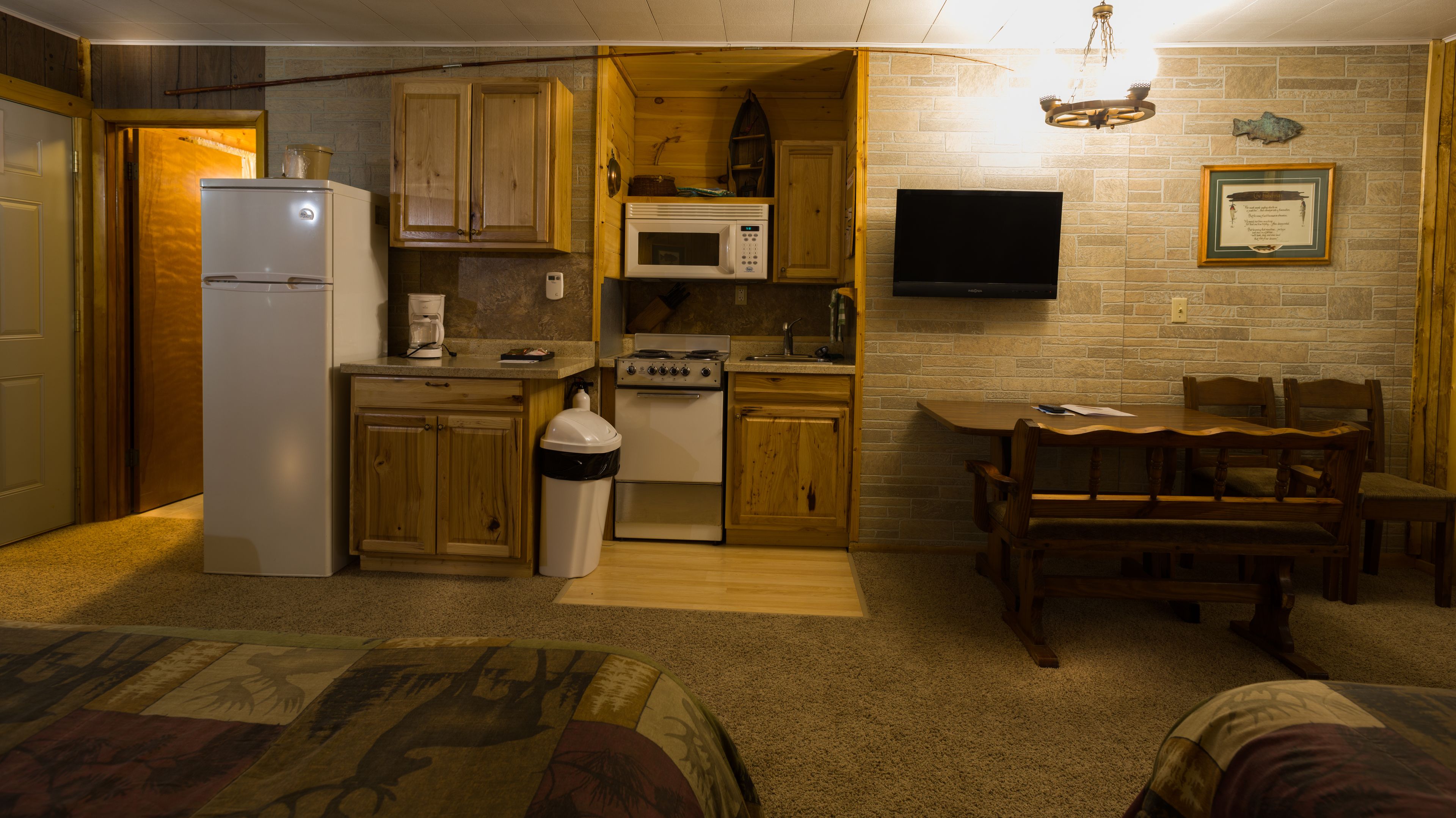 Una cocina compacta con armarios de madera, un microondas, una estufa y un refrigerador. Hay una mesa de comedor con sillas y un televisor montado en la pared.