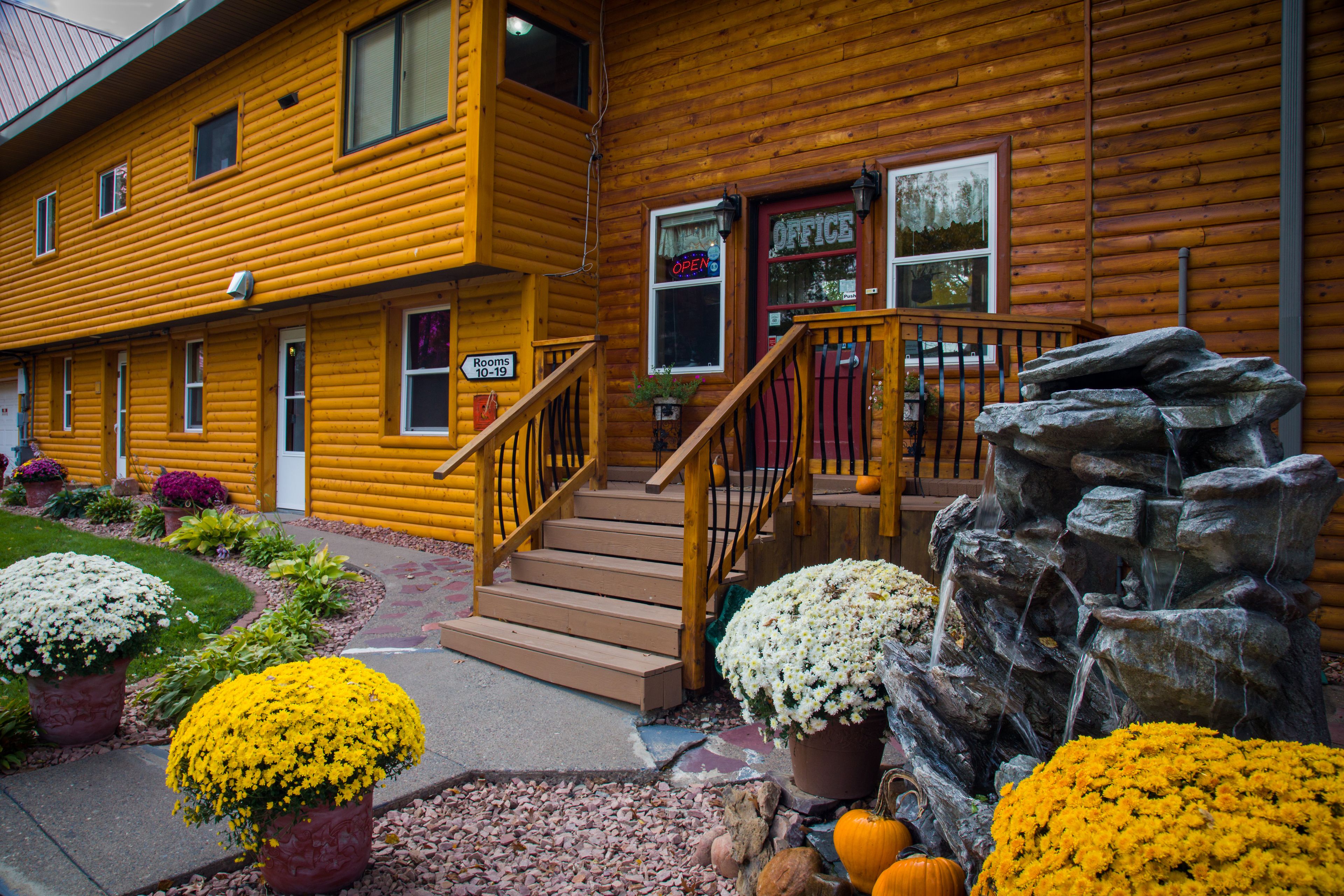 A yellow log cabin with a wooden staircase leading to a porch, surrounded by potted plants and a rock waterfall feature.