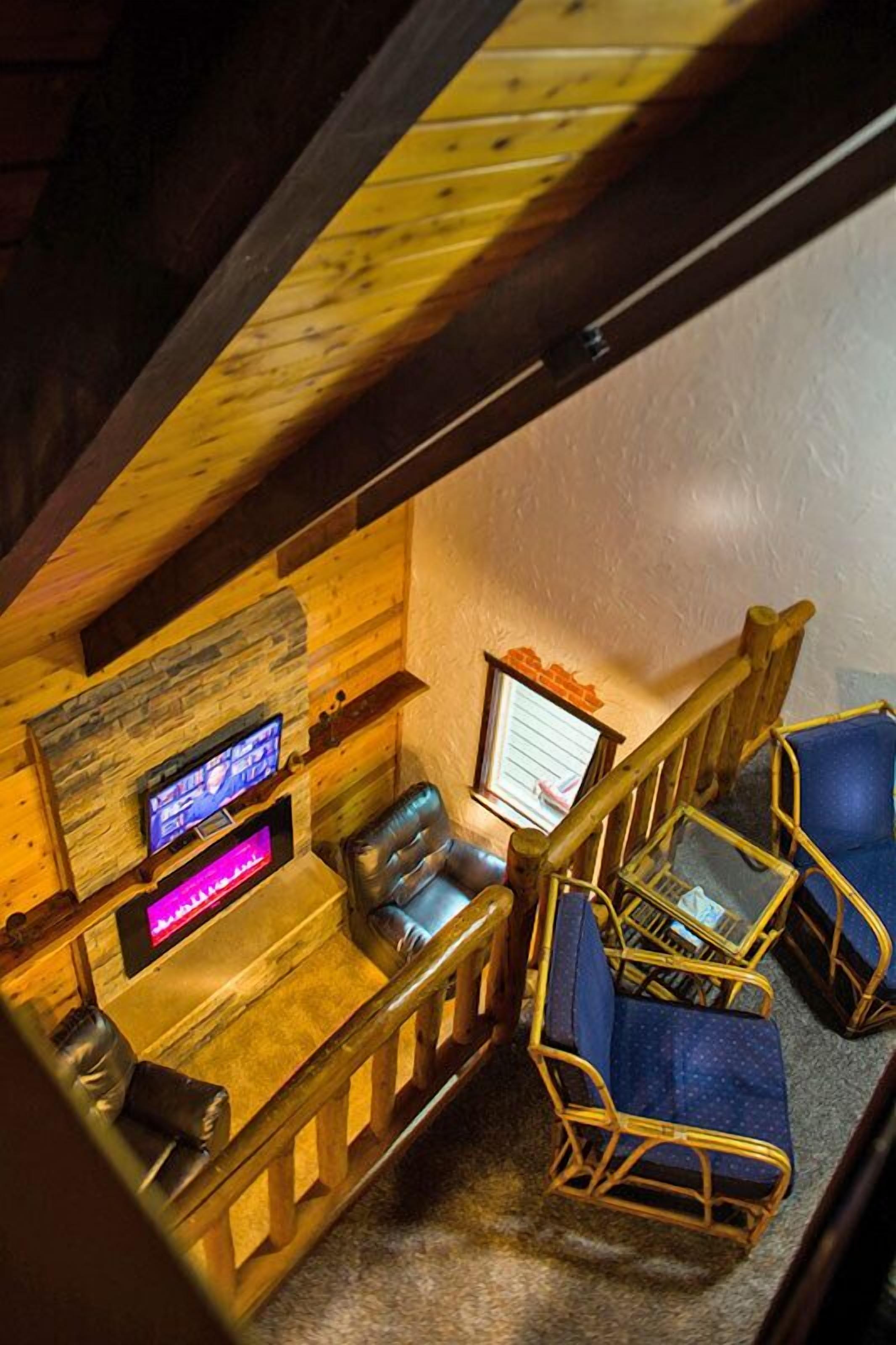 A cozy, rustic room with wooden walls and a sloped ceiling. It features a wooden desk with two chairs, a television, and a small window with a view of the outside.