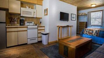 A compact kitchen with a microwave, stove, and sink. There is a wooden table and a couch with a quilt. A TV is mounted on the wall.